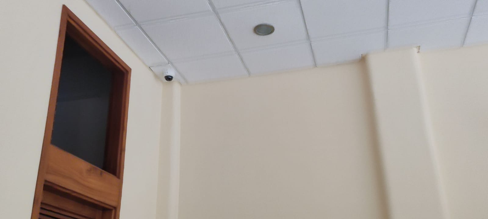 Cámara domo blanca instalada en la esquina de un plafón interior, con cableado por canaleta y una puerta de madera al costado